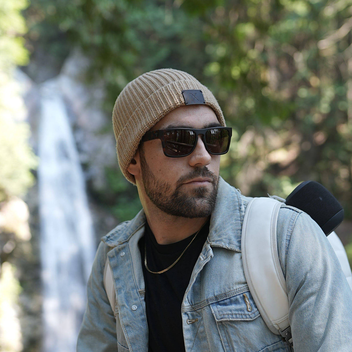 Man wearing Valley Rays sunglasses and a beanie, standing outdoors with a waterfall in the background. Stylish and durable eyewear for outdoor adventures